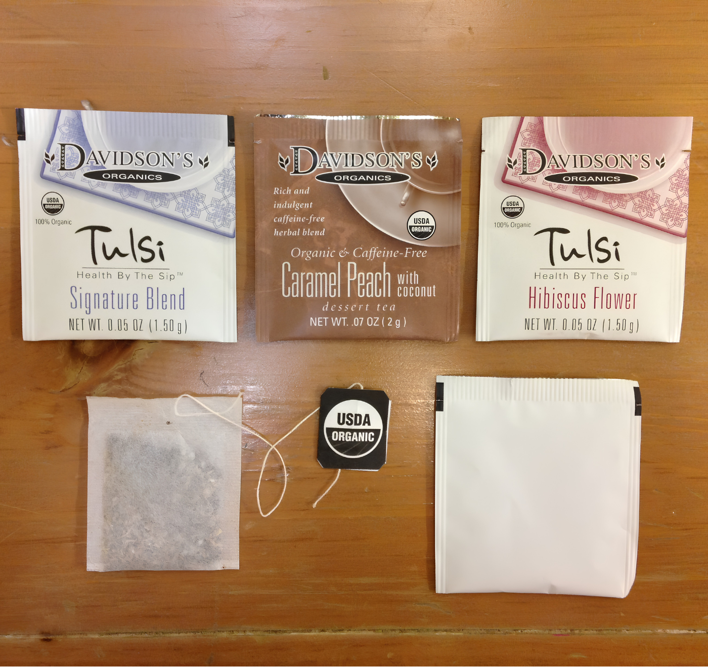 Three Davidson's Organics Tulsi tea bags laid out on a wooden surface, with two white teabags, one brown teabag, and two small tea bags below them.